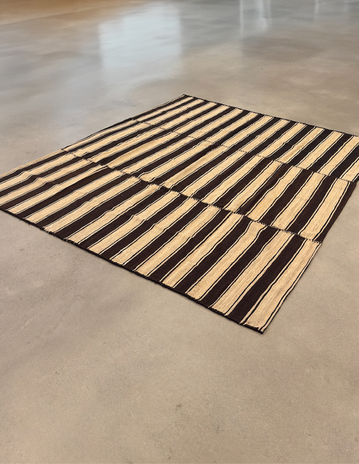 Striped rug with Vintage Iranian Kilim wool rug with a natural beige and brown striped pattern. Handwoven using undyed, raw wool fibers, this one-of-a-kind flatweave textile reflects traditional craftsmanship with a minimal, neutral palette. Its generously scaled 83" × 87" footprint makes it perfect as a centerpiece in living rooms, bedrooms, or creative interiors. The reversible nature and clean-lined geometry make it an ideal choice for mid-century, bohemian, and Japand and beige pattern on a wooden floor