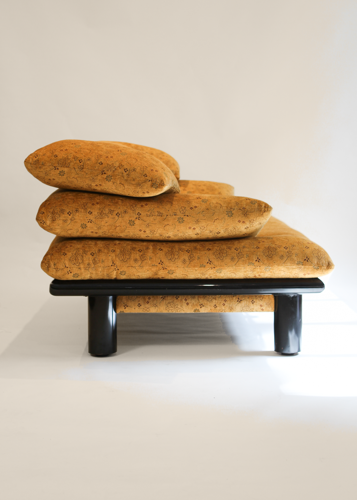 A low black lacquered platform couch with three stacked seat cushions and a matching headrest pillow, photographed against a plain off-white background.